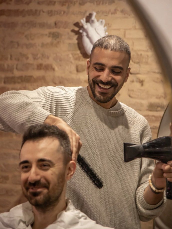 Enrico Cantore ride mentre asciuga i capelli a un cliente con phon e spazzola tonda, nel salone di Via Saragozza